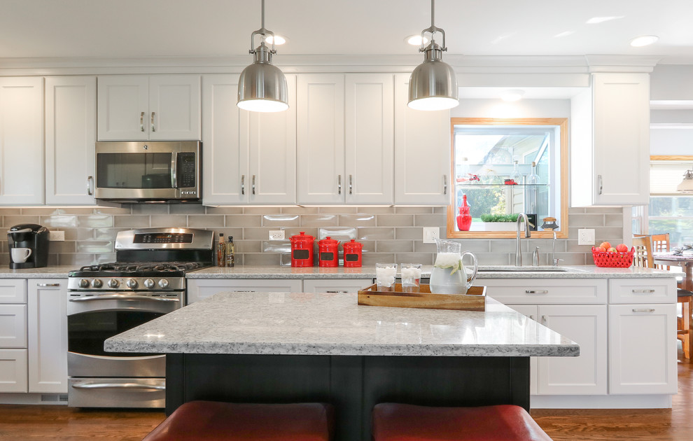 Large traditional galley open plan kitchen in Chicago with a double-bowl sink, shaker cabinets, white cabinets, engineered stone countertops, grey splashback, ceramic splashback, stainless steel appliances, medium hardwood flooring, an island and brown floors.