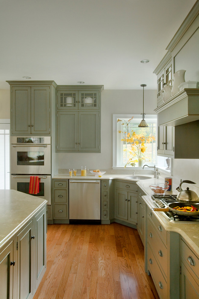 Cette photo montre une cuisine américaine chic en L de taille moyenne avec un placard avec porte à panneau encastré, des portes de placards vertess, un électroménager en acier inoxydable et un sol en bois brun.