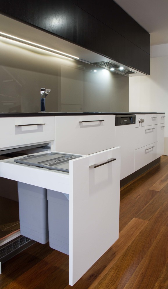 Medium sized modern l-shaped open plan kitchen in Melbourne with flat-panel cabinets, metallic splashback, glass sheet splashback, black appliances, medium hardwood flooring and no island.