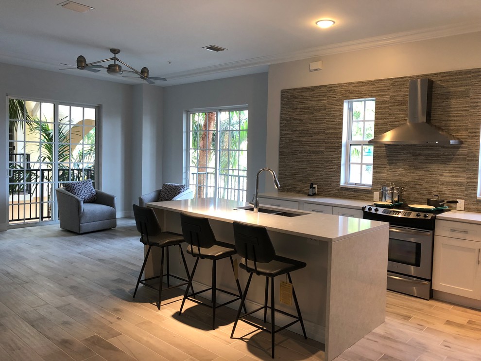 This is an example of a large contemporary l-shaped kitchen/diner in Miami with a submerged sink, shaker cabinets, white cabinets, engineered stone countertops, beige splashback, stone tiled splashback, stainless steel appliances, porcelain flooring, an island and beige floors.