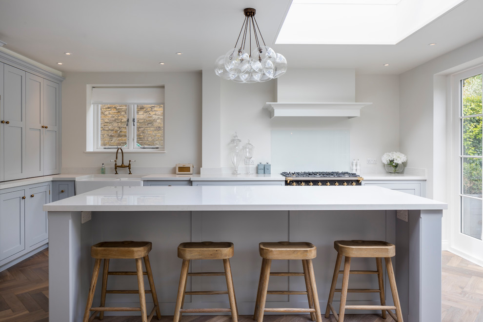 Design ideas for a medium sized contemporary l-shaped kitchen in London with a belfast sink, shaker cabinets, medium hardwood flooring, white splashback, glass sheet splashback, black appliances and an island.