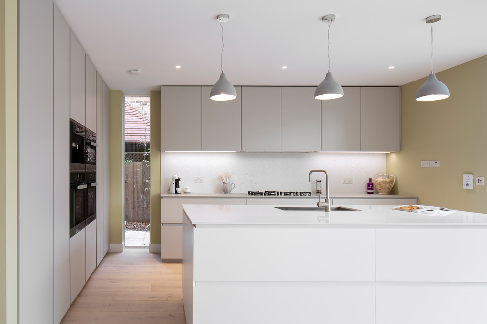 Offene, Mittelgroße Moderne Küche mit Waschbecken, flächenbündigen Schrankfronten, beigen Schränken, Quarzit-Arbeitsplatte, Küchenrückwand in Weiß, Rückwand aus Stein, schwarzen Elektrogeräten, hellem Holzboden und Kücheninsel in Oxfordshire