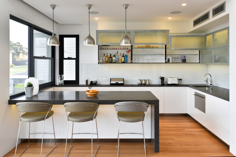 Moderne Küche in L-Form mit Unterbauwaschbecken, flächenbündigen Schrankfronten, weißen Schränken, Küchenrückwand in Weiß, Glasrückwand, weißen Elektrogeräten, braunem Holzboden, Halbinsel und braunem Boden in Brisbane