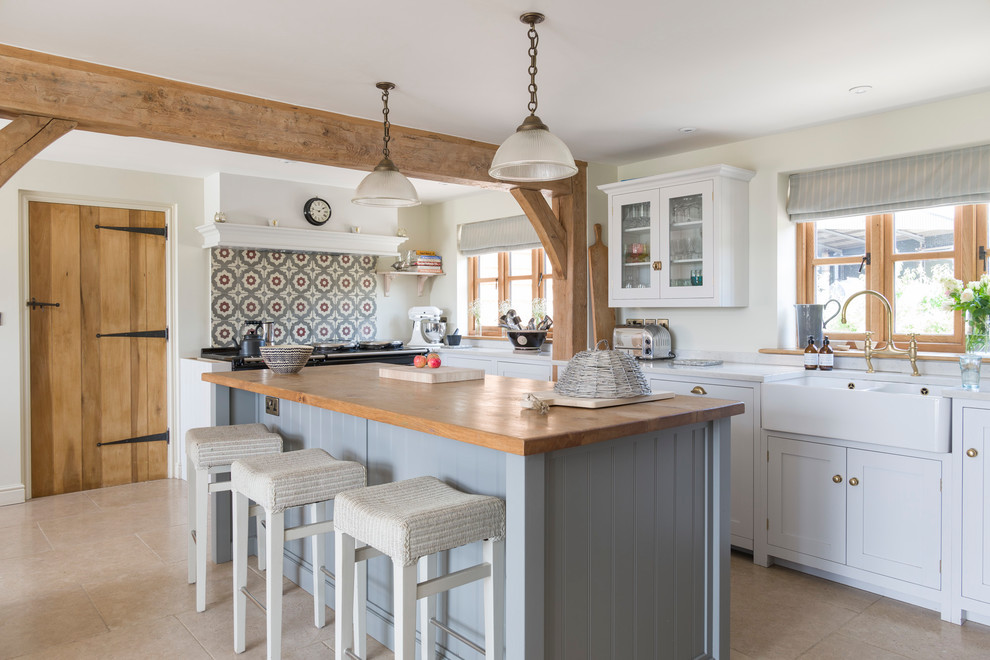 Rural l-shaped kitchen in West Midlands with a belfast sink, shaker cabinets, white cabinets, multi-coloured splashback, an island, beige floors and white worktops.