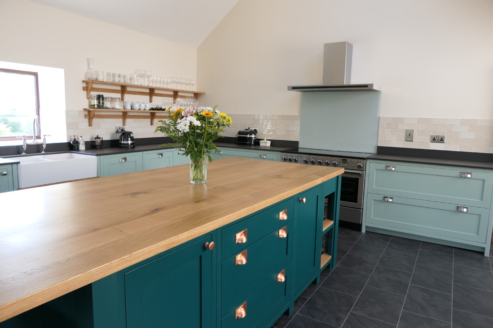 Large classic l-shaped open plan kitchen in West Midlands with a belfast sink, shaker cabinets, green cabinets, engineered stone countertops, grey splashback, glass sheet splashback, stainless steel appliances, ceramic flooring, an island, black floors and black worktops.