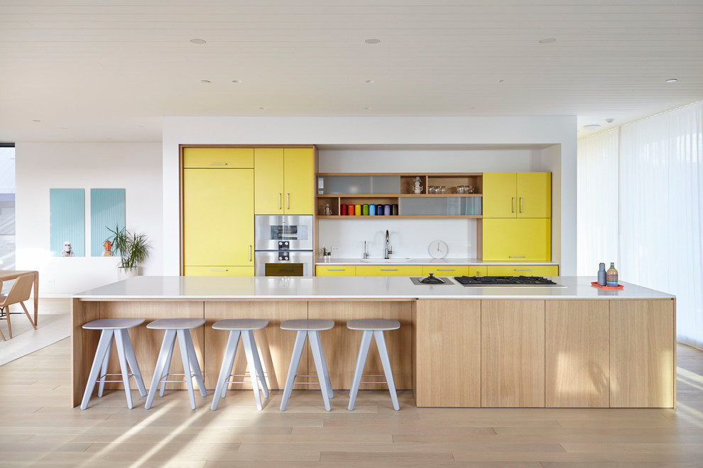 Mid-sized trendy galley light wood floor and beige floor open concept kitchen photo in Los Angeles with flat-panel cabinets, yellow cabinets, white backsplash, stainless steel appliances, an island, white countertops, an undermount sink and solid surface countertops