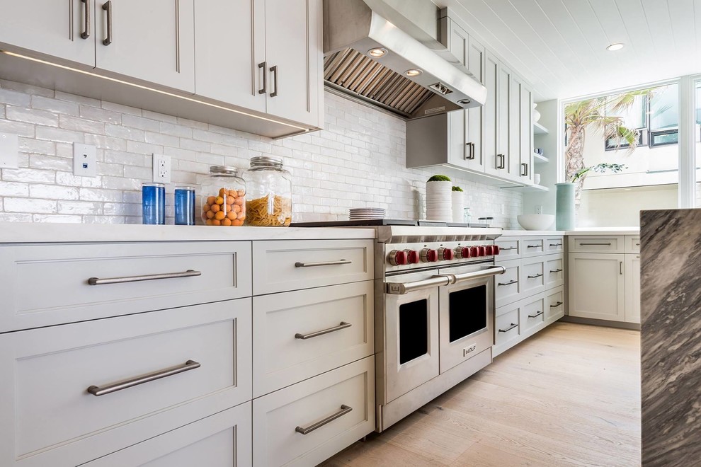 Kitchen - mid-sized coastal l-shaped light wood floor and beige floor kitchen idea in Los Angeles with shaker cabinets, gray cabinets, quartz countertops, white backsplash, ceramic backsplash, stainless steel appliances, an island and white countertops