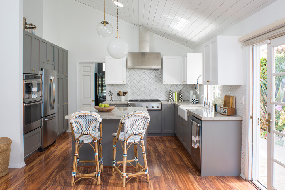Photo of a medium sized farmhouse grey and white u-shaped kitchen in Los Angeles with a belfast sink, shaker cabinets, grey cabinets, engineered stone countertops, white splashback, porcelain splashback, stainless steel appliances, medium hardwood flooring and an island.