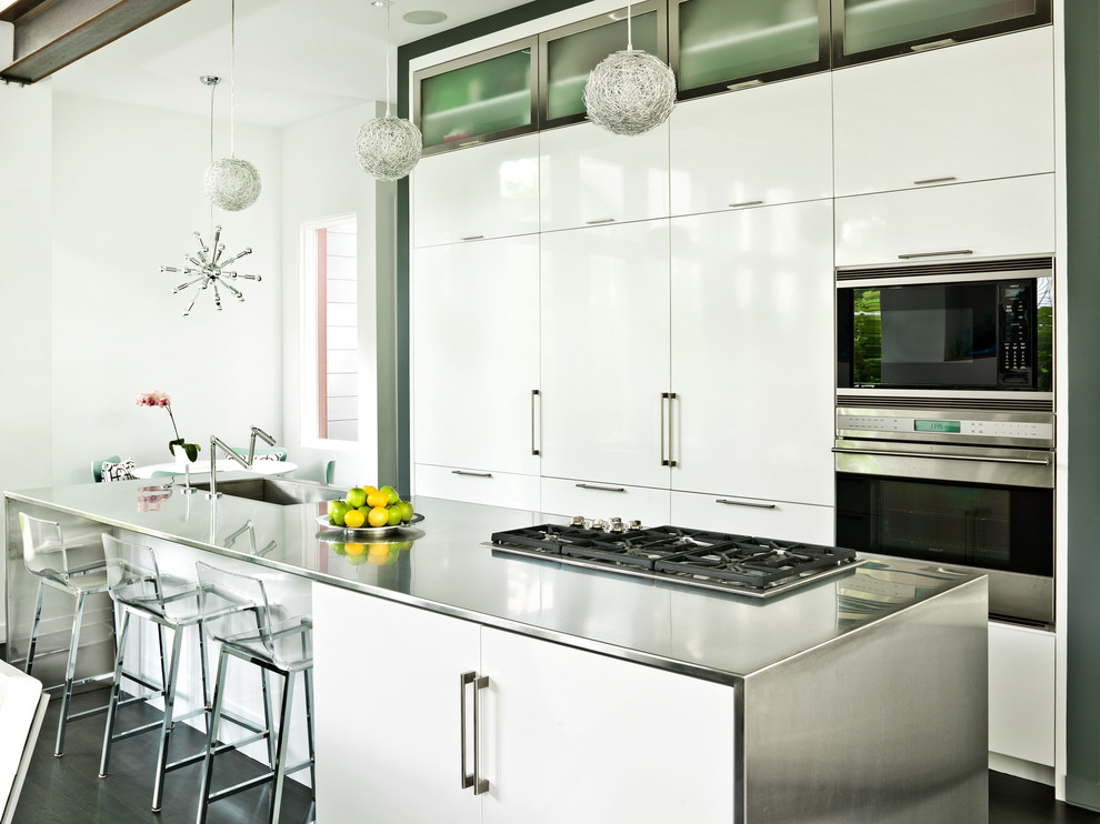 Cette image montre une cuisine américaine parallèle design avec un évier intégré, un placard à porte plane, des portes de placard blanches, un plan de travail en inox et un électroménager en acier inoxydable.