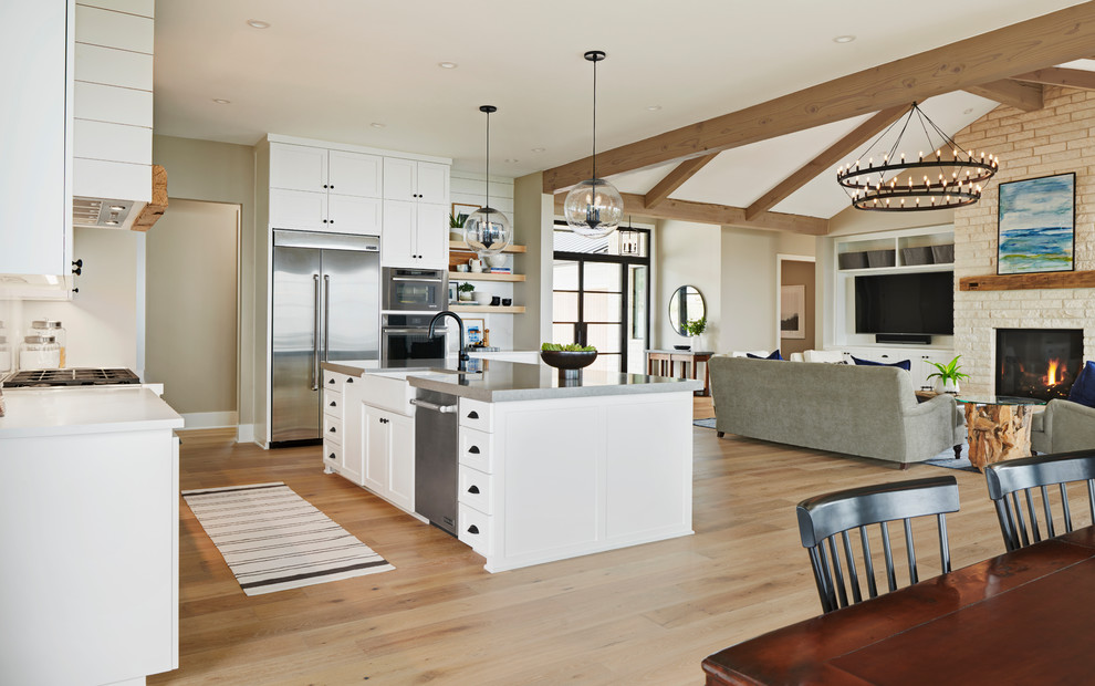 Inspiration for a large rural l-shaped open plan kitchen in Austin with a belfast sink, shaker cabinets, white cabinets, engineered stone countertops, white splashback, stone slab splashback, stainless steel appliances, light hardwood flooring, an island, brown floors, white worktops and a feature wall.