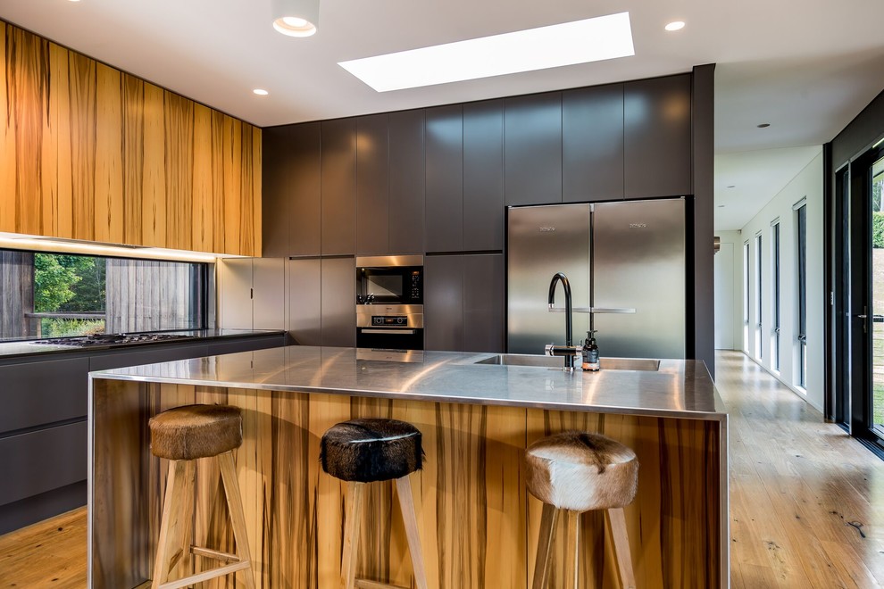 Moderne Küche in L-Form mit integriertem Waschbecken, flächenbündigen Schrankfronten, grauen Schränken, Edelstahl-Arbeitsplatte, Küchengeräten aus Edelstahl, braunem Holzboden, Kücheninsel und braunem Boden in Wollongong