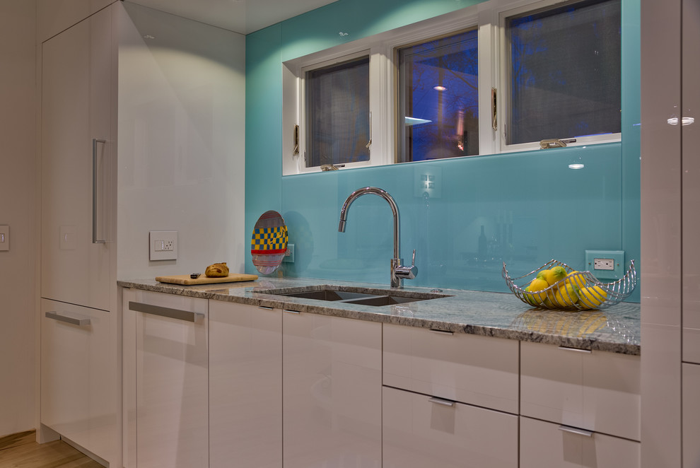 Bild på ett mellanstort funkis kök, med en undermonterad diskho, släta luckor, vita skåp, granitbänkskiva, blått stänkskydd, vita vitvaror, ljust trägolv och en köksö