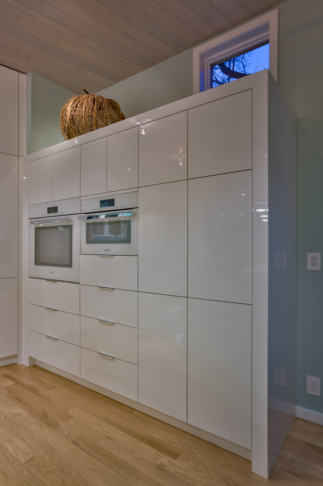 This is an example of a medium sized contemporary l-shaped kitchen/diner in Omaha with a submerged sink, flat-panel cabinets, white cabinets, granite worktops, blue splashback, white appliances, light hardwood flooring and an island.