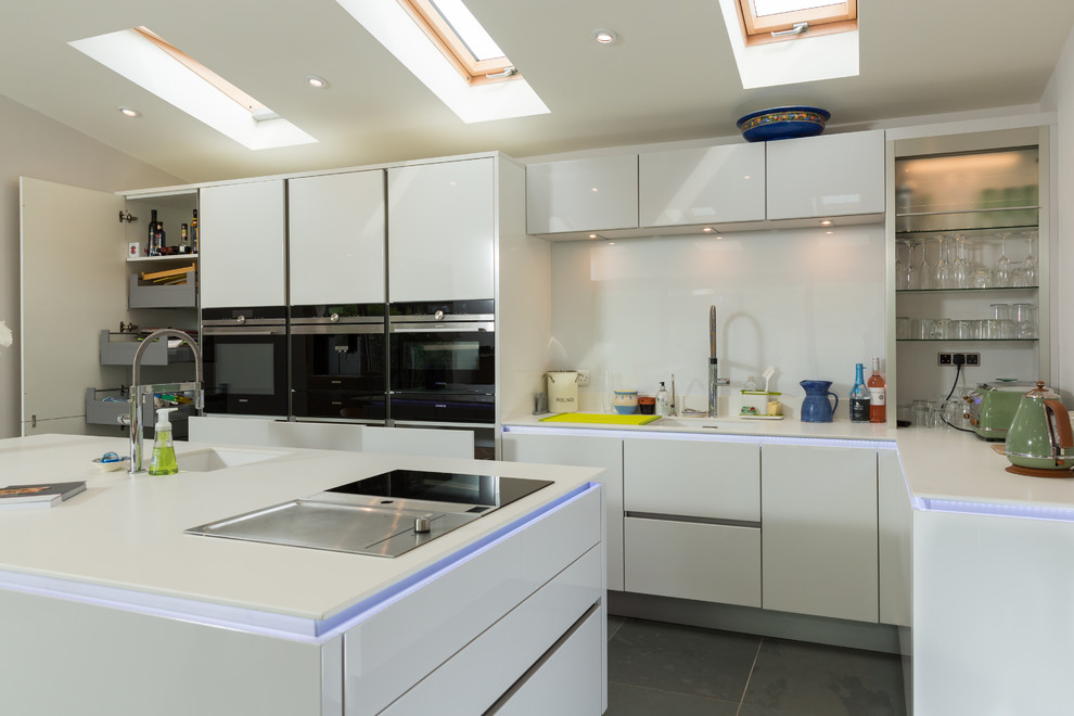 Inspiration for a large contemporary single-wall open plan kitchen in Hampshire with an integrated sink, flat-panel cabinets, white cabinets, composite countertops, white splashback, glass sheet splashback, stainless steel appliances, ceramic flooring and an island.