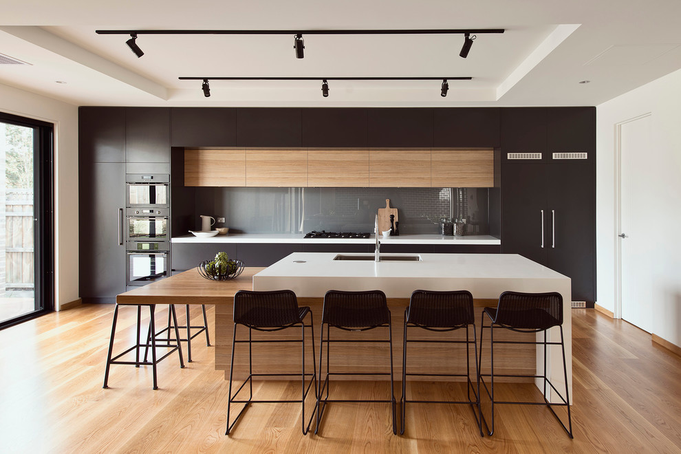 Zweizeilige, Große Moderne Wohnküche mit Unterbauwaschbecken, flächenbündigen Schrankfronten, schwarzen Schränken, Glasrückwand, schwarzen Elektrogeräten, Kücheninsel und braunem Holzboden in Melbourne