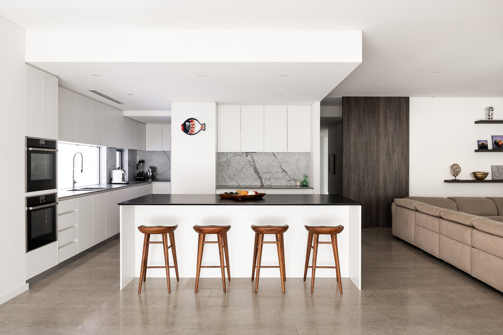 Trendy l-shaped beige floor open concept kitchen photo in Perth with flat-panel cabinets, white cabinets, gray backsplash, black appliances, an island and black countertops