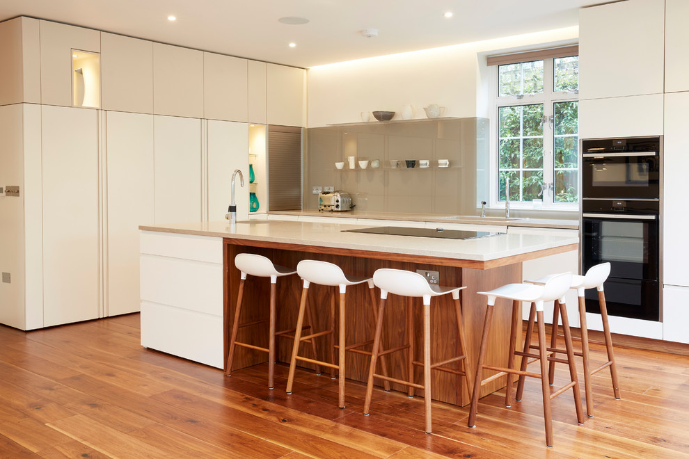 Moderne Küche mit flächenbündigen Schrankfronten, weißen Schränken, Küchenrückwand in Beige, Glasrückwand, schwarzen Elektrogeräten, braunem Holzboden und Kücheninsel in London