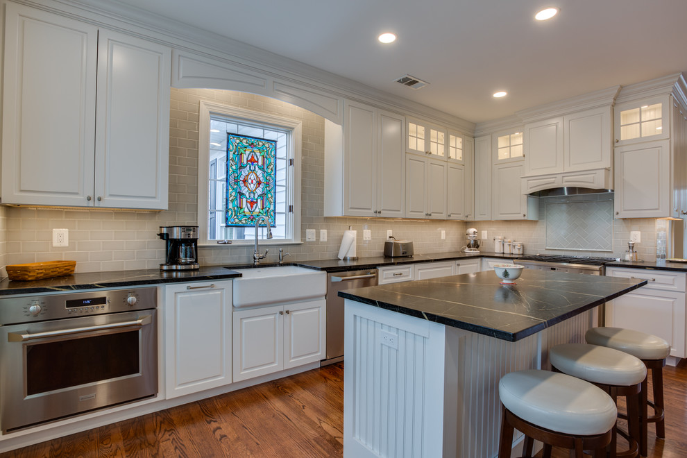 This is an example of a classic l-shaped enclosed kitchen in Baltimore with a belfast sink, raised-panel cabinets, white cabinets, grey splashback, metro tiled splashback, stainless steel appliances, medium hardwood flooring, an island and brown floors.