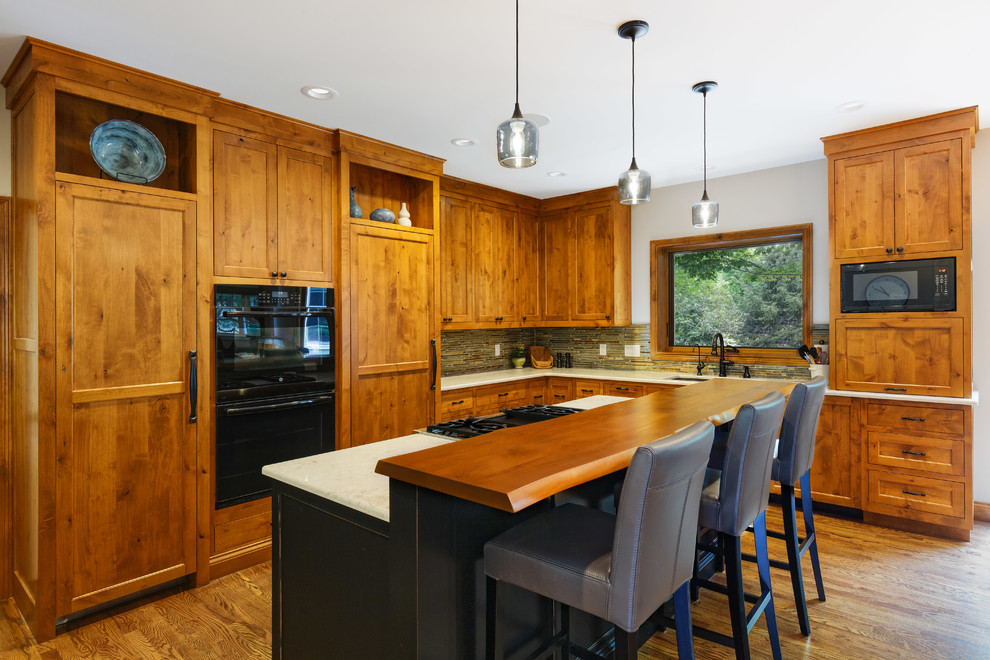 Cette photo montre une cuisine américaine montagne en bois brun avec un placard à porte plane, un plan de travail en granite, une crédence marron, un électroménager noir, un sol en bois brun et îlot.