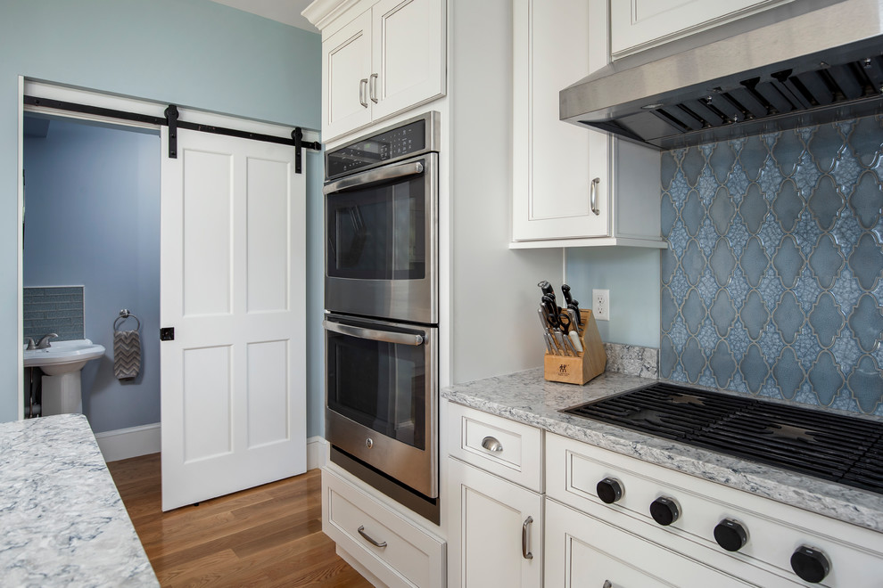 Example of a mid-sized transitional l-shaped medium tone wood floor kitchen pantry design in Boston with a farmhouse sink, flat-panel cabinets, white cabinets, quartz countertops, blue backsplash, ceramic backsplash, stainless steel appliances, an island and white countertops