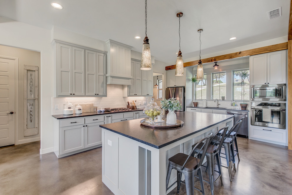 Example of a mid-sized country galley concrete floor eat-in kitchen design in Dallas with a farmhouse sink, shaker cabinets, gray cabinets, white backsplash, an island and black countertops