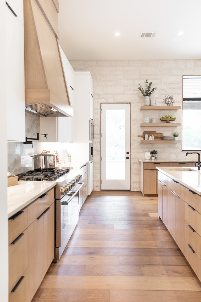 Inspiration for an expansive modern l-shaped open plan kitchen in Austin with flat-panel cabinets, light wood cabinets, quartz worktops, stainless steel appliances, an island, a belfast sink, light hardwood flooring and brown floors.