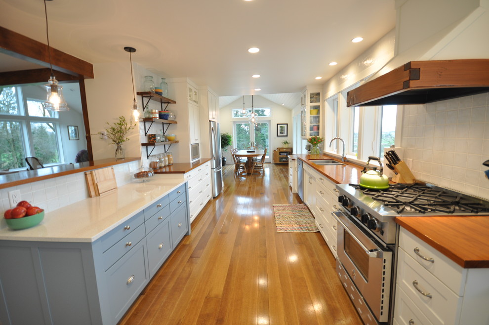 This is an example of a large contemporary galley kitchen/diner in Vancouver with a submerged sink, shaker cabinets, white cabinets, wood worktops, white splashback, porcelain splashback, stainless steel appliances, medium hardwood flooring and a breakfast bar.