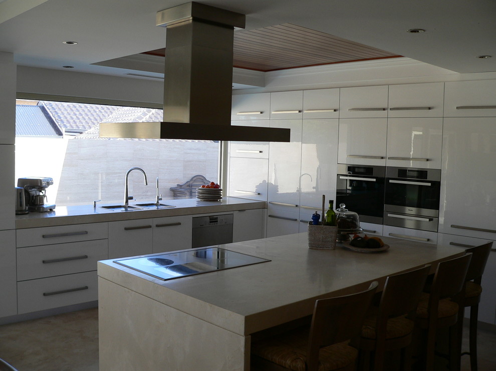 Maritim inredning av ett stort kök, med en undermonterad diskho, vita skåp, marmorbänkskiva, svarta vitvaror, travertin golv och en köksö