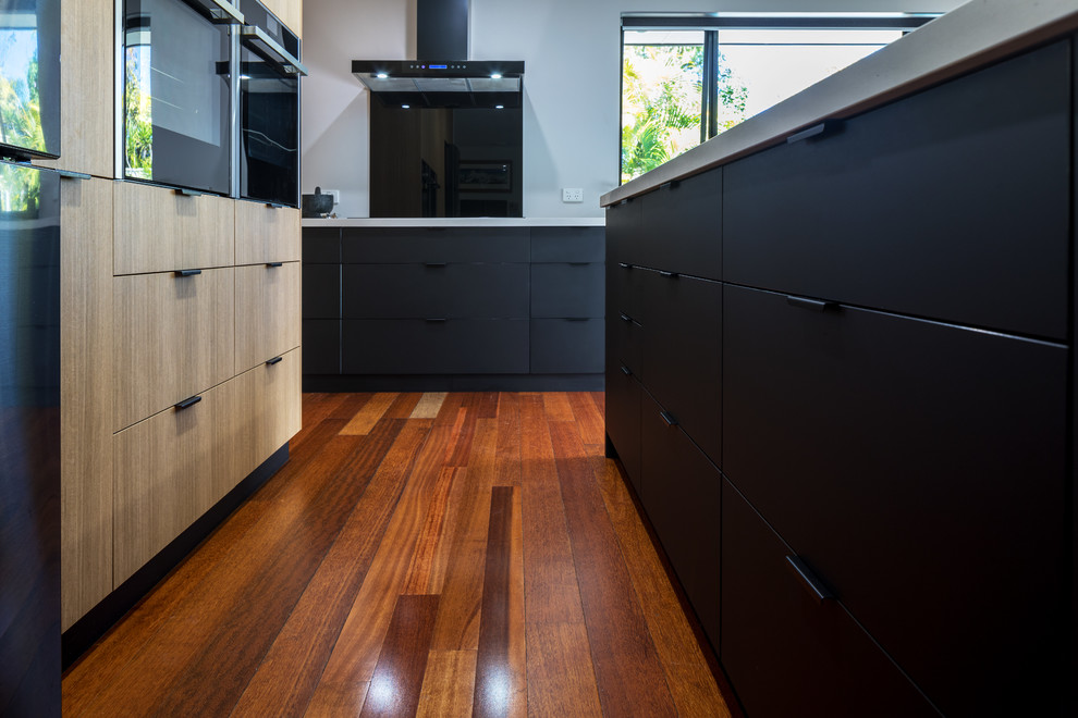 Exemple d'une cuisine moderne en bois clair avec un placard à porte plane, un plan de travail en quartz modifié, une crédence noire, une crédence en feuille de verre, un électroménager noir, îlot et un plan de travail gris.