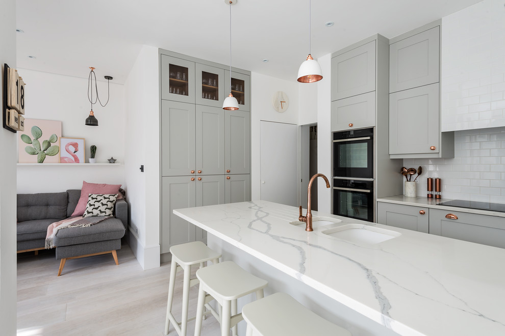 This is an example of a medium sized contemporary l-shaped open plan kitchen in London with an integrated sink, shaker cabinets, grey cabinets, quartz worktops, white splashback, metro tiled splashback, stainless steel appliances, light hardwood flooring, an island and grey floors.