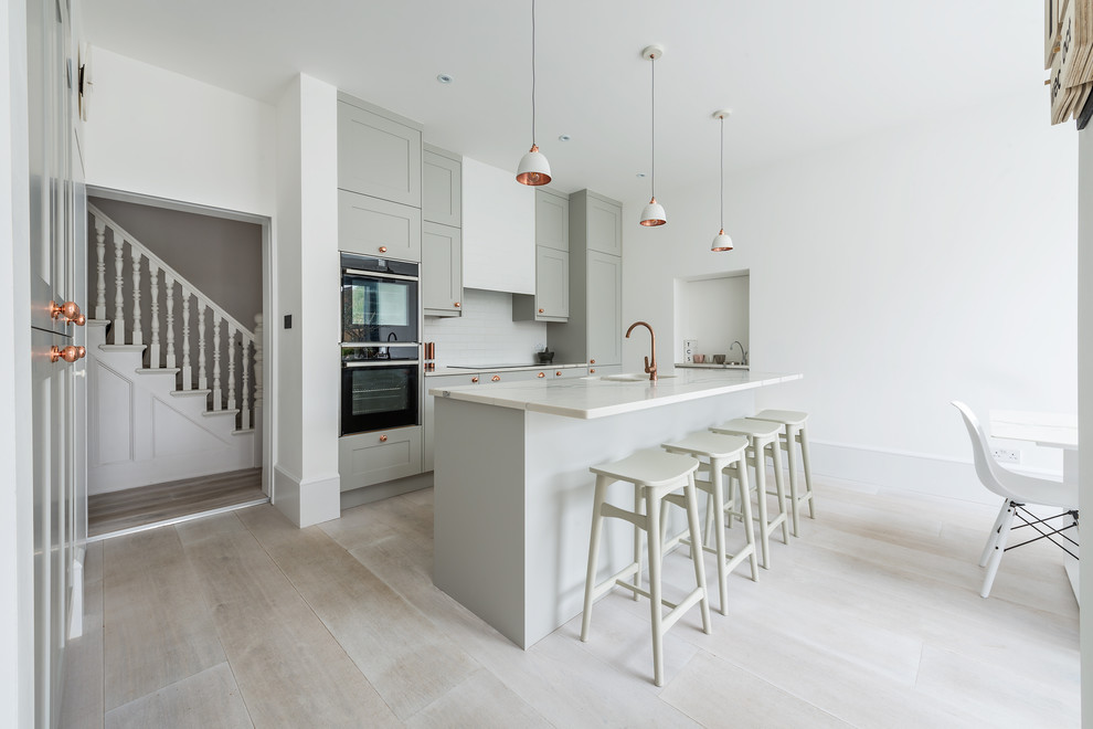 Inspiration for a medium sized contemporary l-shaped open plan kitchen in London with an integrated sink, shaker cabinets, grey cabinets, quartz worktops, white splashback, metro tiled splashback, stainless steel appliances, light hardwood flooring, an island and grey floors.