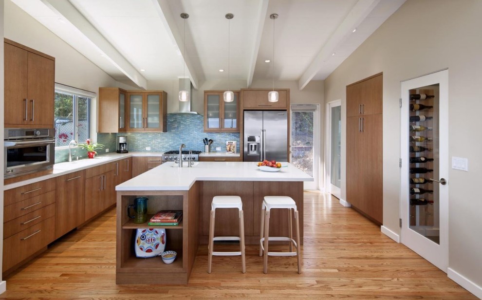 Mittelgroße Moderne Küche in L-Form mit flächenbündigen Schrankfronten, hellbraunen Holzschränken, Küchenrückwand in Blau, Rückwand aus Glasfliesen, Küchengeräten aus Edelstahl, hellem Holzboden, Kücheninsel, braunem Boden und weißer Arbeitsplatte in Denver