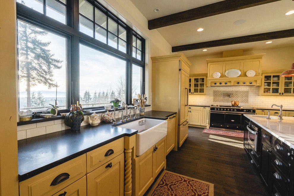 Example of a large transitional l-shaped medium tone wood floor and brown floor open concept kitchen design in Seattle with a farmhouse sink, raised-panel cabinets, yellow cabinets, soapstone countertops, white backsplash, subway tile backsplash, black appliances, an island and gray countertops