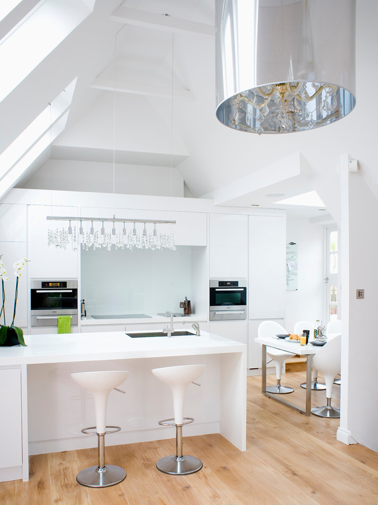 Bild på ett mellanstort funkis vit vitt kök och matrum, med en undermonterad diskho, släta luckor, vita skåp, glaspanel som stänkskydd, rostfria vitvaror, ljust trägolv, en köksö och beiget golv