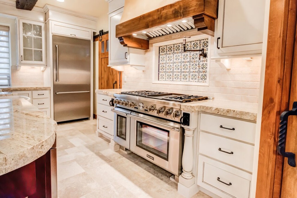 Example of a mid-sized mountain style travertine floor eat-in kitchen design in Orange County with a farmhouse sink, beaded inset cabinets, white cabinets, granite countertops, beige backsplash, stone tile backsplash, stainless steel appliances and an island