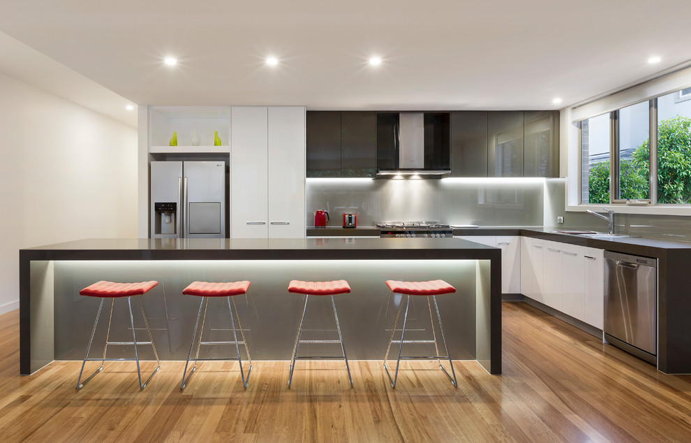 Moderne Wohnküche in L-Form mit Unterbauwaschbecken, flächenbündigen Schrankfronten, weißen Schränken, Küchenrückwand in Grau, Glasrückwand, Küchengeräten aus Edelstahl, braunem Holzboden, Kücheninsel, braunem Boden und grauer Arbeitsplatte in Melbourne