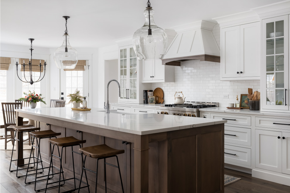 Bild på ett lantligt vit vitt kök och matrum, med vita skåp, vitt stänkskydd, stänkskydd i tunnelbanekakel, rostfria vitvaror, mörkt trägolv, en köksö, brunt golv och skåp i shakerstil