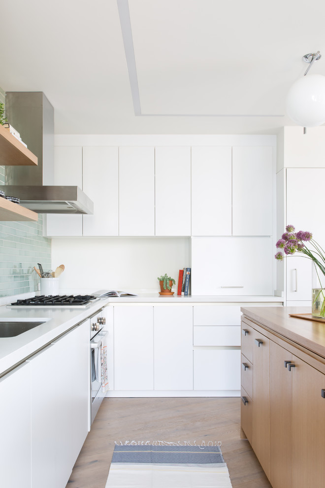 Retro inredning av ett vit vitt kök, med släta luckor, vita skåp, grönt stänkskydd, rostfria vitvaror, ljust trägolv, en köksö och beiget golv