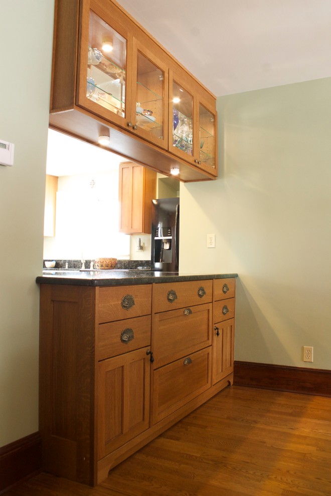 Example of a mid-sized arts and crafts l-shaped medium tone wood floor eat-in kitchen design in Detroit with a farmhouse sink, glass-front cabinets, medium tone wood cabinets, granite countertops and black appliances