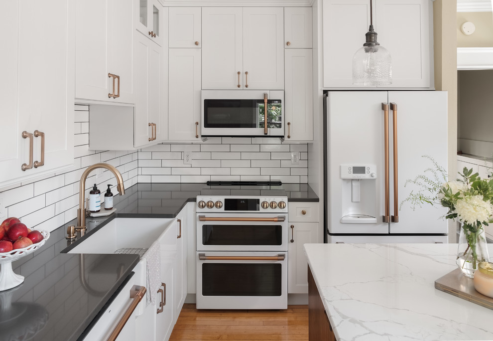 Mid-sized elegant l-shaped light wood floor eat-in kitchen photo in Other with a farmhouse sink, shaker cabinets, white cabinets, quartz countertops, white backsplash, ceramic backsplash, white appliances, an island and gray countertops