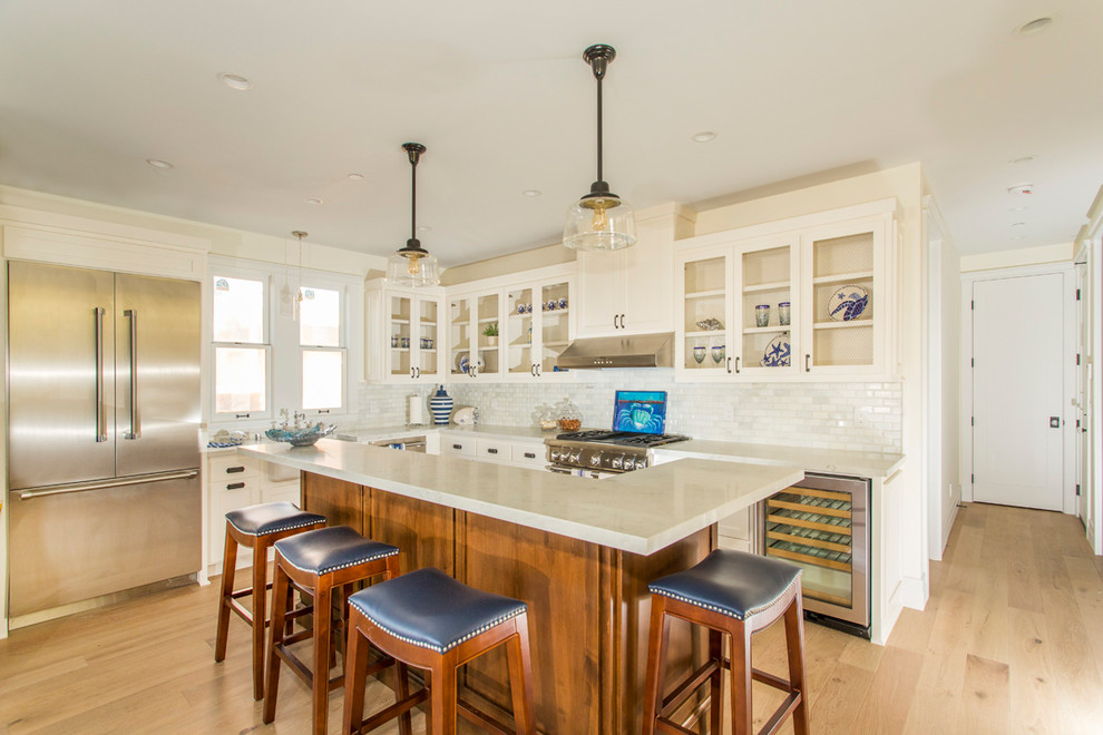 Beach style l-shaped medium tone wood floor kitchen photo in San Diego with white cabinets, white backsplash, stainless steel appliances and an island