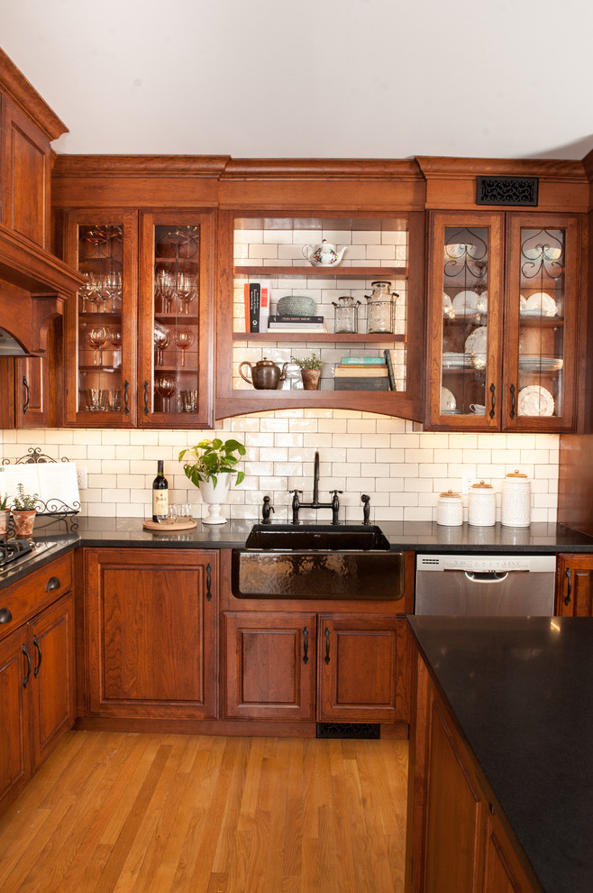 Inspiration for a large timeless l-shaped medium tone wood floor and brown floor eat-in kitchen remodel in Providence with a farmhouse sink, glass-front cabinets, medium tone wood cabinets, solid surface countertops, white backsplash, subway tile backsplash, stainless steel appliances, an island and black countertops