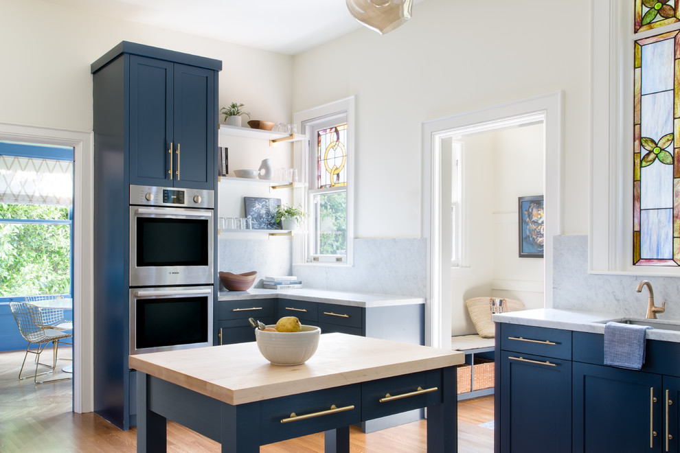 Example of a transitional u-shaped medium tone wood floor and brown floor enclosed kitchen design in San Francisco with blue cabinets, marble backsplash, stainless steel appliances, an island, an undermount sink, shaker cabinets, marble countertops, gray backsplash and gray countertops