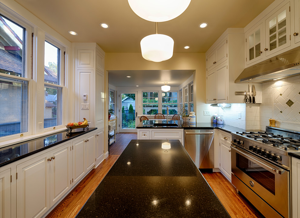 This is an example of a medium sized traditional galley kitchen/diner in Seattle with a built-in sink, raised-panel cabinets, white cabinets, granite worktops, white splashback, metro tiled splashback, stainless steel appliances, light hardwood flooring, an island and beige floors.