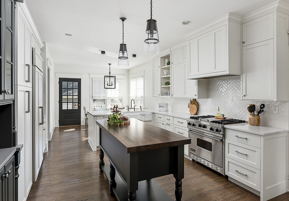 This is an example of a medium sized classic u-shaped kitchen/diner in Chicago with a belfast sink, shaker cabinets, white cabinets, engineered stone countertops, white splashback, porcelain splashback, stainless steel appliances, dark hardwood flooring, a breakfast bar and brown floors.