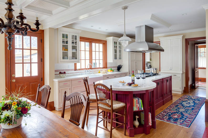 Klassische Wohnküche mit Landhausspüle, Kassettenfronten, roten Schränken, Marmor-Arbeitsplatte, Küchenrückwand in Weiß, Rückwand aus Stein, Elektrogeräten mit Frontblende, braunem Holzboden und Kücheninsel in Boston