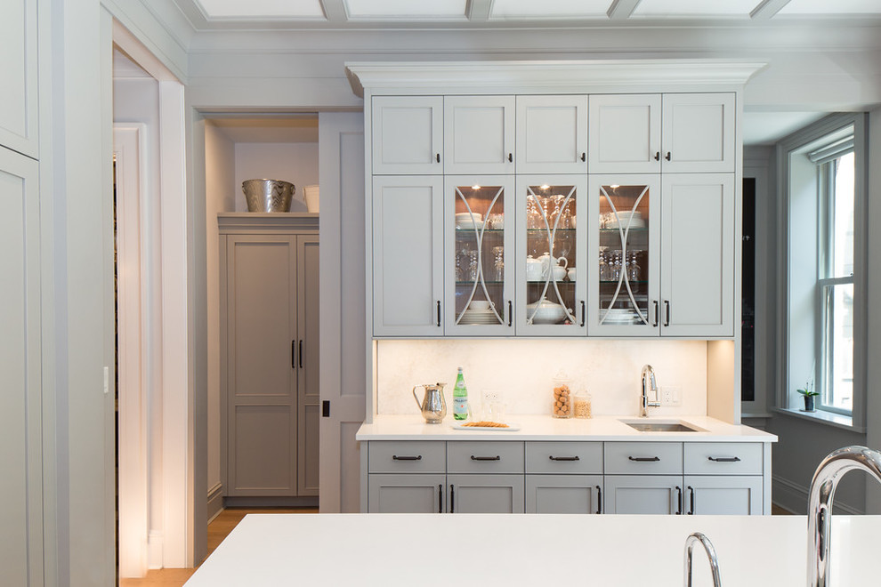 Inredning av ett klassiskt avskilt kök, med en undermonterad diskho, luckor med infälld panel, grå skåp, bänkskiva i kvarts, vitt stänkskydd, integrerade vitvaror, ljust trägolv och en köksö