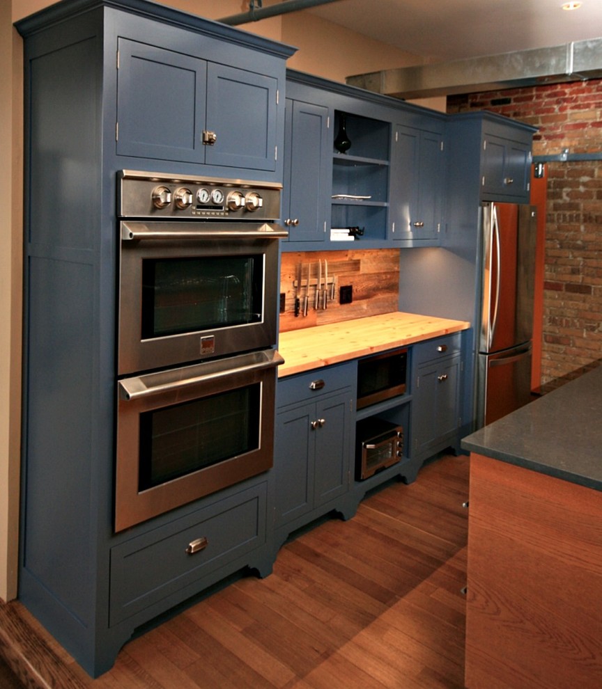 Diseño de cocina rústica con armarios estilo shaker, puertas de armario azules, encimera de madera, salpicadero marrón, electrodomésticos de acero inoxidable, suelo de madera en tonos medios y una isla