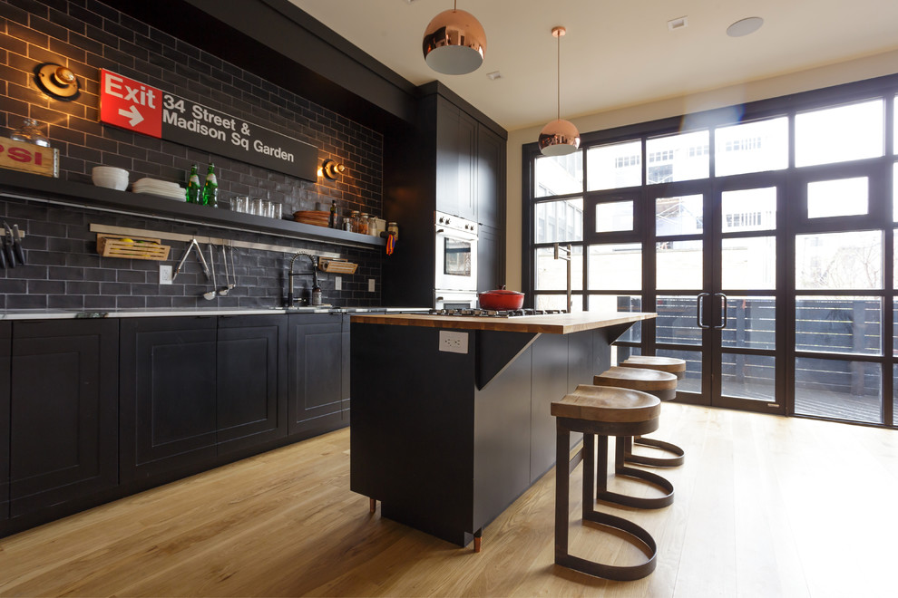 Bild på ett mellanstort industriellt kök, med en undermonterad diskho, skåp i shakerstil, svarta skåp, träbänkskiva, grått stänkskydd, stänkskydd i tunnelbanekakel, rostfria vitvaror, ljust trägolv och en köksö