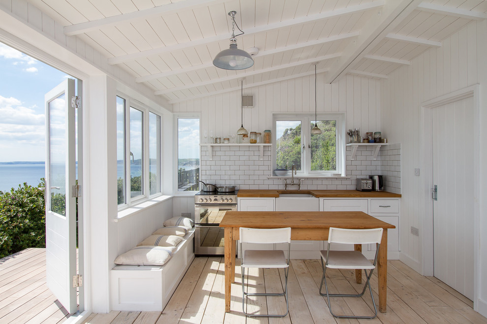 Example of a mid-sized beach style single-wall light wood floor eat-in kitchen design in Cornwall with a farmhouse sink, white cabinets, white backsplash, subway tile backsplash, stainless steel appliances, shaker cabinets and wood countertops